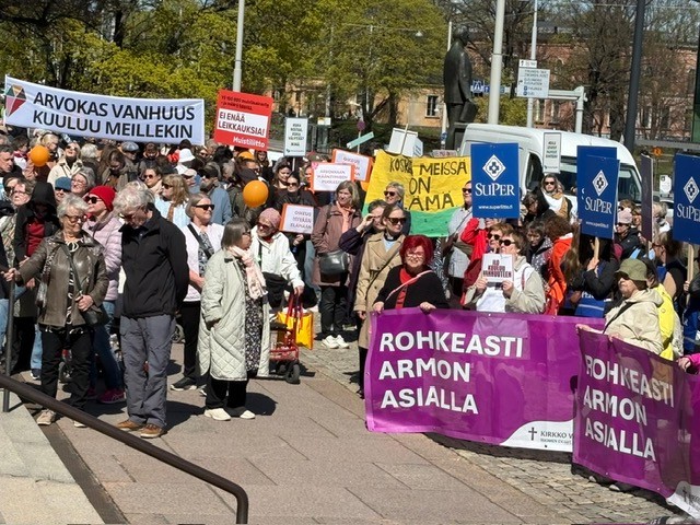 Rollaattorimarssin osallistujia kokoontuneena kadulle Helsingissä. Ihmiset pitävät käsissään värikkäitä kylttejä ja banderolleja, joissa viestitään hyvän ja arvokkaan vanhuuden puolesta. Kuvassa näkyy eri-ikäisiä osallistujia aurinkoisessa säässä sekä järjestöjen tunnuksia ja tapahtuman yhteisöllinen tunnelma.