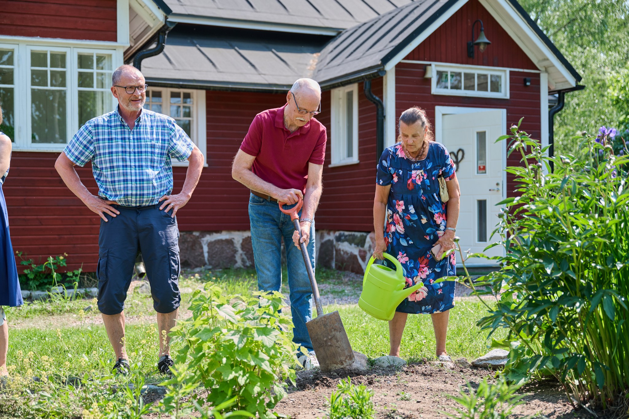 Kolme iäkästä ihmistä hoitaa yhdessä pihaa aurinkoisena kesäpäivänä. Yksi kaivaa maata lapiolla, toinen kastelee istutuksia vihreällä kastelukannulla ja kolmas seuraa vieressä työn etenemistä. Taustalla näkyy punainen puutalo ja vehreä piha. Kuvassa välittyvät yhteisöllisyys, aktiivinen arki, yhdessä tekeminen ja omasta elinympäristöstä huolehtiminen.