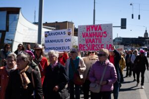 Iäkkäitä ihmisiä marssii kaupungin keskustassa kylttien kanssa. Kylteissä lukee ”Arvokas vanhuus on ihmisoikeus” ja ”Hyvä vanhuus vaatii resursseja”.
