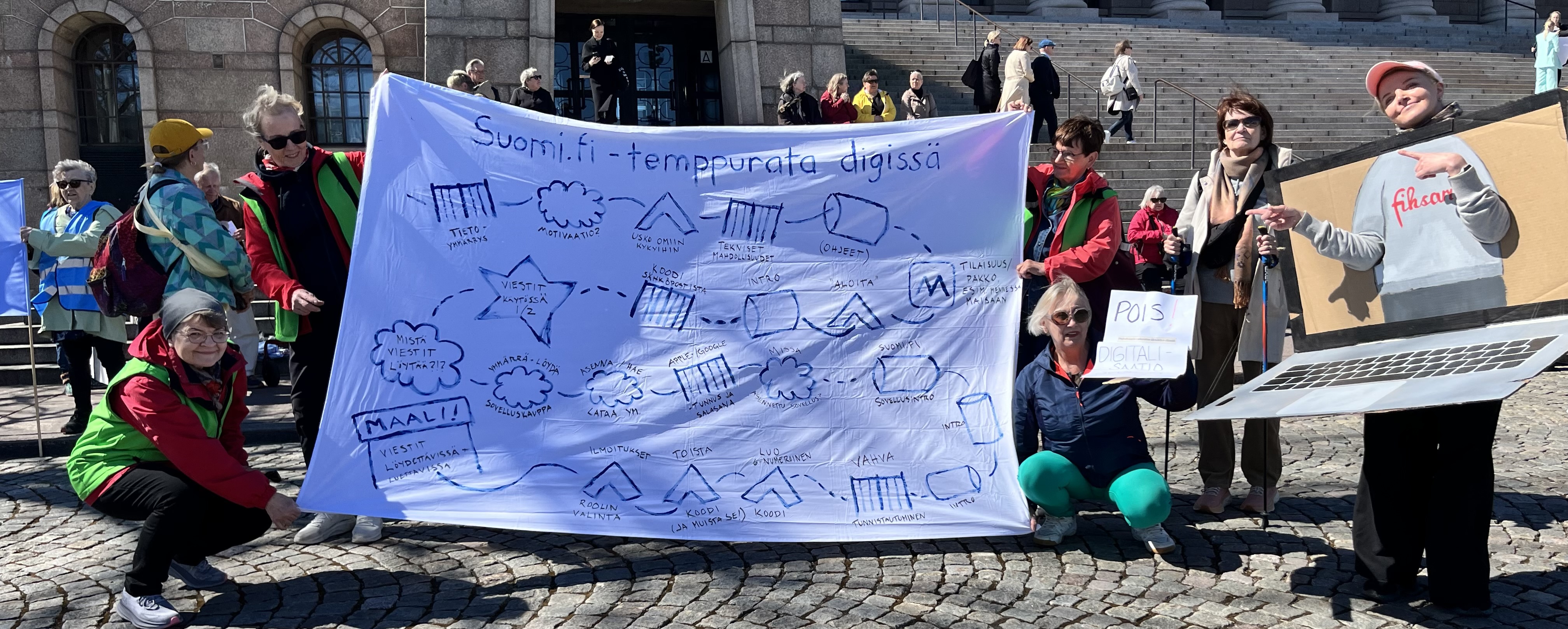 Ryhmä ihmisiä seisoo Eduskuntatalon portaiden edessä osallistumassa Rollaattorimarssiin. He pitelevät suurta kangasta, johon on piirretty "Suomi.fi-temppurata digissä" sekä digipalvelujen käyttöä kuvaava reitti esteineen ja vaiheineen. Oikealla yksi osallistuja on pukeutunut suureksi kannettavaksi tietokoneeksi, jonka näytöllä näkyy "Fiksari"-hahmo. Vieressä toinen osallistuja pitää kylttiä, jossa lukee: "Pois digitaalista syrjäytystä". Tunnelma on näkyvä ja vaikuttava, ja kuva korostaa ikääntyneiden kokemuksia digipalvelujen haasteista sekä tarvetta saavutettavammille palveluille.