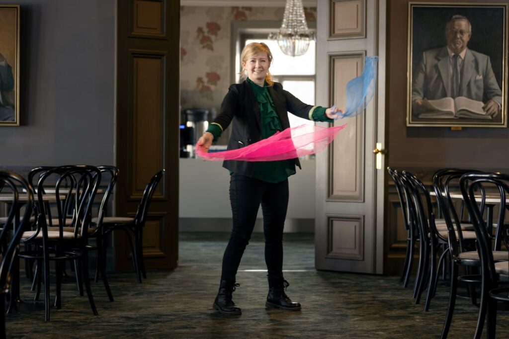 Hymyilevä nainen seisoo juhlavassa salissa ja liikuttaa käsissään pinkkiä ja sinistä huivia. Ympärillä on pöytiä ja tuoleja, ja seinällä näkyy suuri muotokuva.