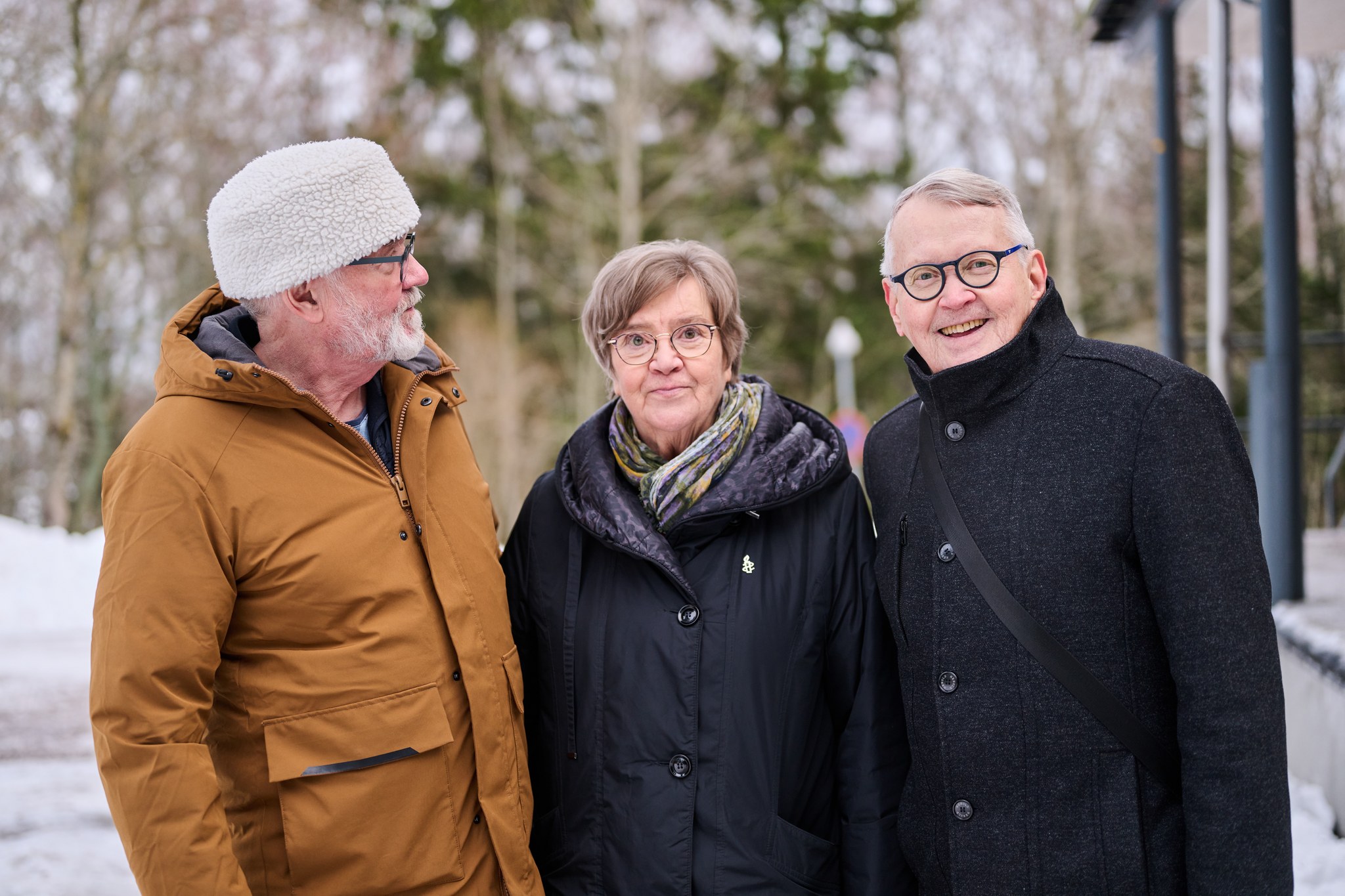 Kolme iäkästä ihmistä seisoo vierekkäin ulkona talvisessa puistomaisemassa. Vasemmalla valkopartainen mies ruskeassa takissa ja valkoisessa pipossa katsoo keskellä seisovaa silmälasipäistä naista. Oikealla tummaan takkiin pukeutunut, silmälasipäinen mies hymyilee kameralle.