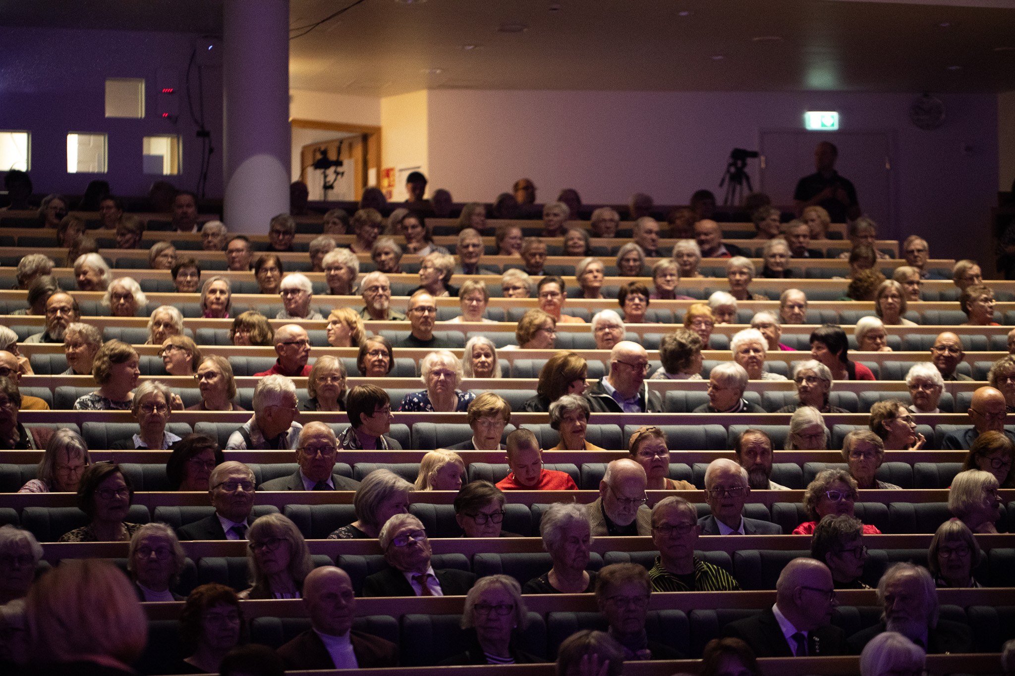 Suuri joukko ihmisiä istuu auditoriossa odottamassa tilaisuuden alkua. Yleisössä on runsaasti iäkkäitä osallistujia, jotka keskustelevat keskenään.