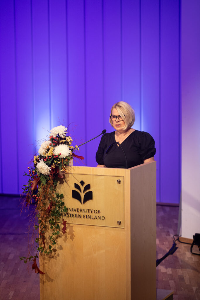 Nainen pitää puhetta kukin koristellun puhujakorokkeen takana. Taustalla on siniviolettina valaistu juhlasali.