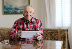 Hymyilevä iäkäs mies istuu pöydän ääressä ja käyttää tablettia. Hänellä on yllään punainen ruutupaita, ja pöydällä on silmälasit.