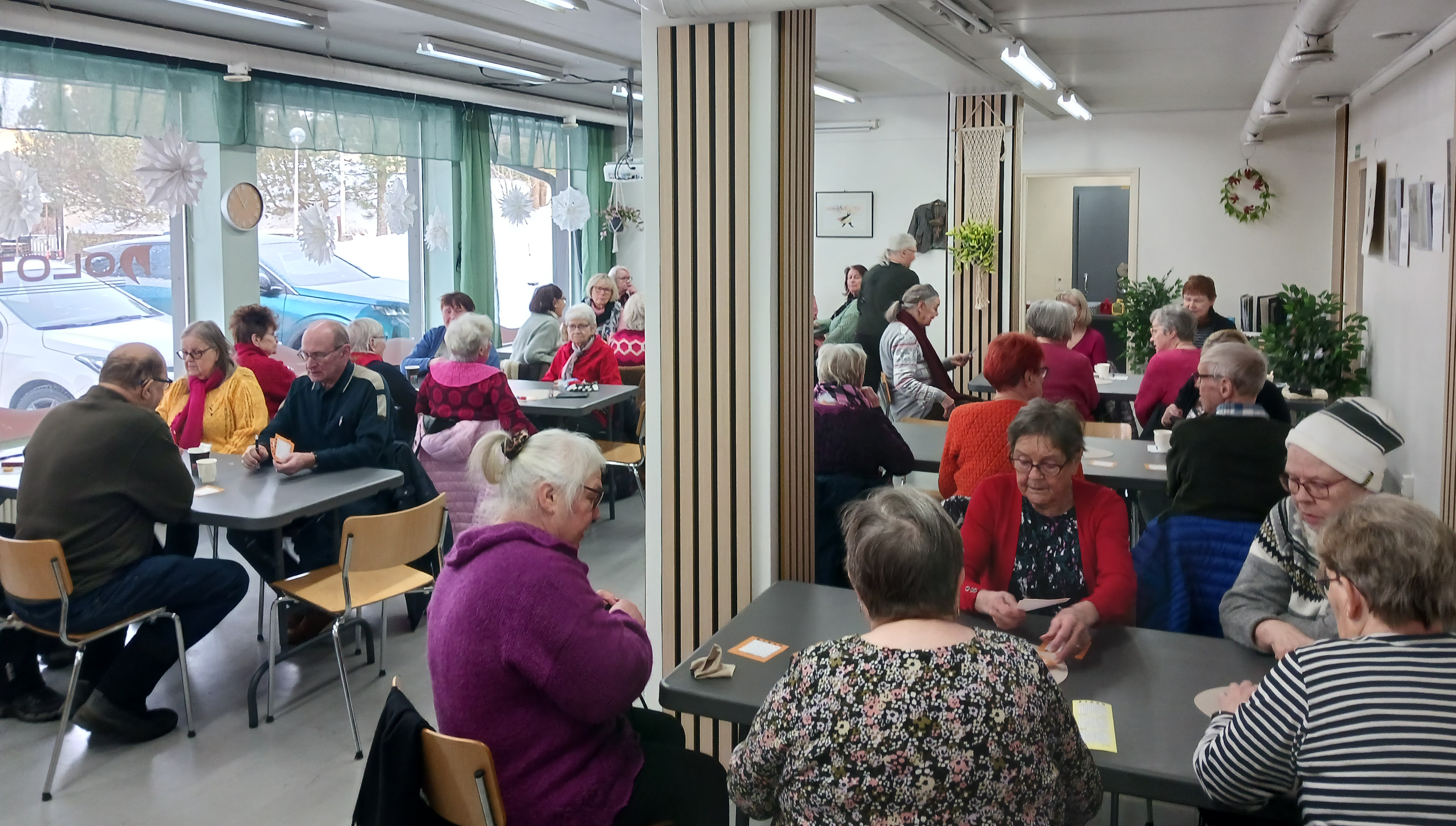 Iäkkäitä ihmisiä istuu pienissä ryhmissä pöytien ääressä valoisassa kohtaamispaikassa. Osallistujat keskustelevat keskenään ja viettävät aikaa yhdessä.